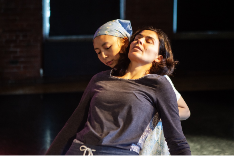 A woman (Maria Kapsali) leans back into a dark room. She is leaning on another woman, who holds her back. Both of their faces are visible with eyes downcast in a restful gaze.