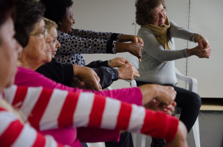 Five older women stretch their arms forward on front of their bodies while they are sitting down