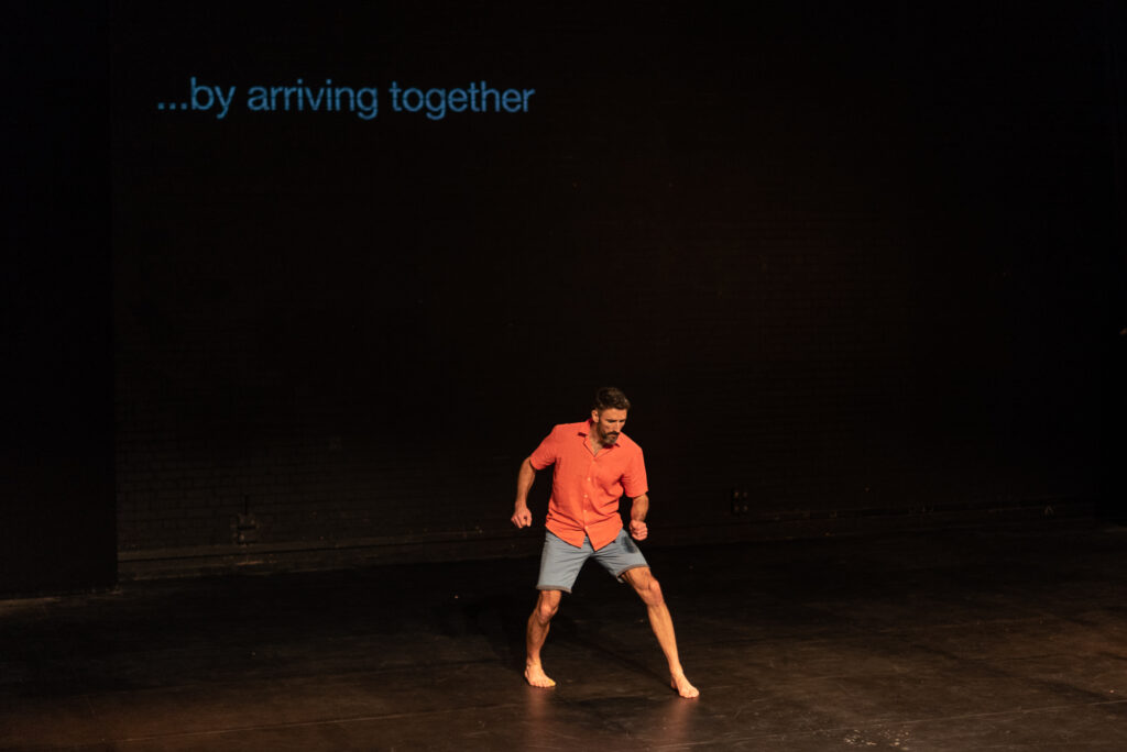 A man stands in the foreground of a stage with a red shirt and blue shorts, his left leg is stepping forward and right leg back, his arms are bent. On a screen in the background it states '...by arriving together'.